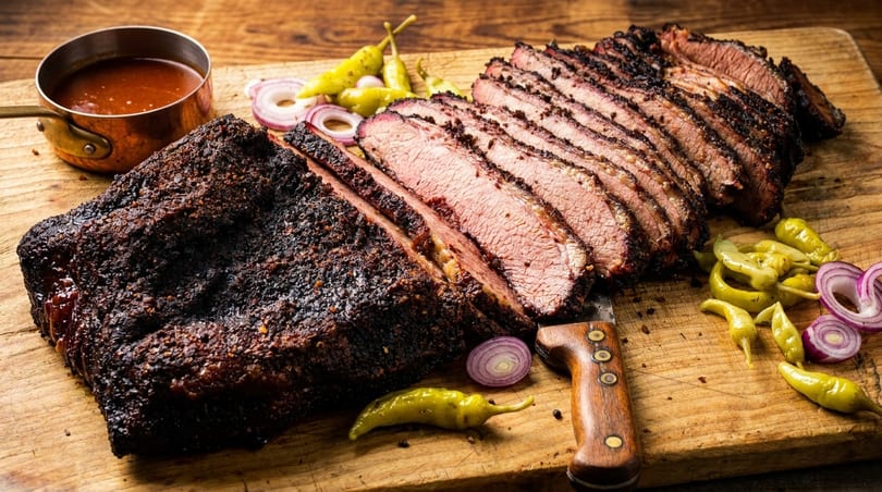 Sliced smoked brisket with a dark crust, served with sauce, pickled peppers, and red onion.