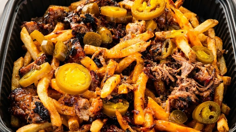 Close-up of loaded fries with pulled pork, barbecue sauce, and sliced jalapeños in a black container.