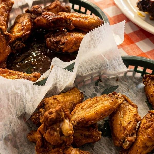 Two baskets of crispy chicken wings, one with sauce, on a red and white checkered tablecloth.