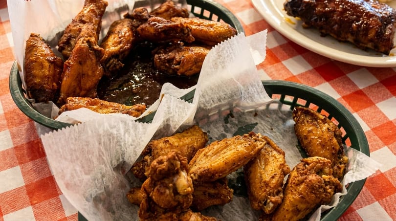 Two baskets of crispy chicken wings, one with sauce, on a red and white checkered tablecloth.